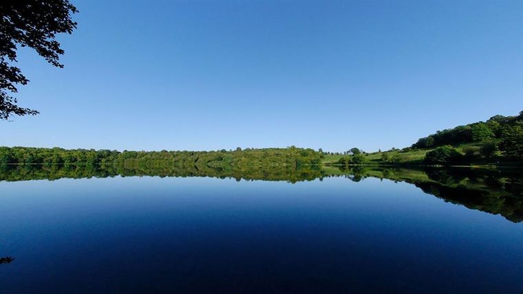 Ein ruhiger See mit klarem Wasser und spiegelndem Himmel. Umgeben von üppigem Grün und einem blauen, wolkenlosen Himmel.