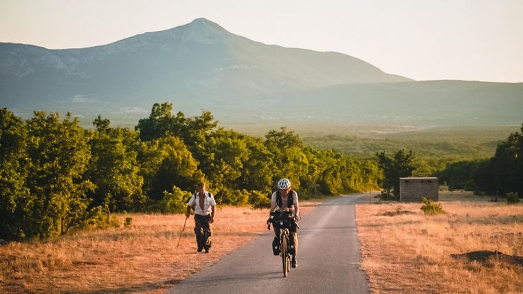 Zwei Personen sind auf einem Naturweg unterwegs. Eine Person wandert, während die andere mit dem Fahrrad fährt, umgeben von einer schönen Landschaft und Bergen im Hintergrund.