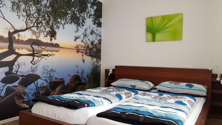 Een gezellige slaapkamer met een tweepersoonsbed en een groot muurschilderij van een rustige meer. Aan de muur hangt een groen schilderij met een plant.