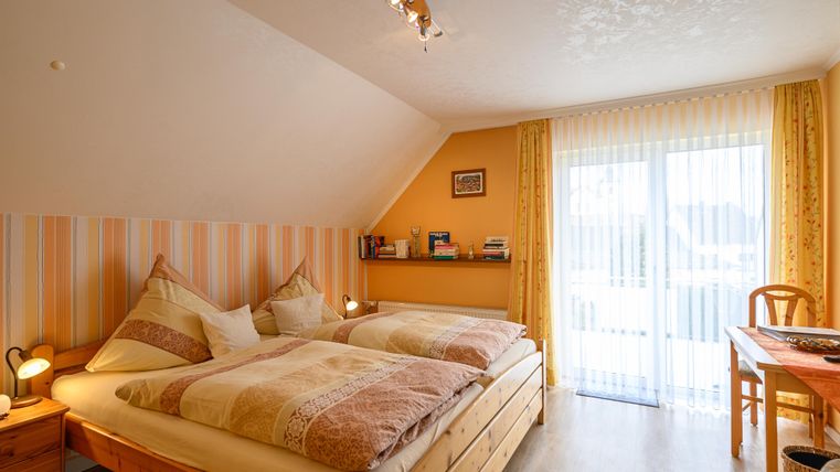 Ein gemütliches Schlafzimmer mit zwei Betten und warmen Farbtonen. Große Fenster lassen viel Licht herein und bieten Zugang zu einem Balkon.