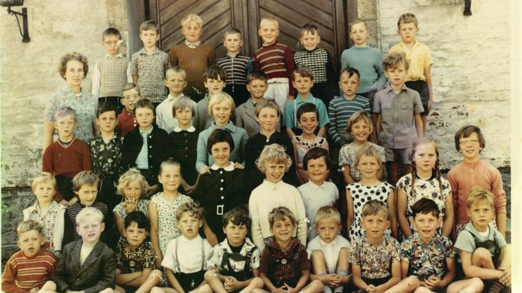 A group of children is standing in front of a large door. They are dressed in a variety of styles and are smiling at the camera.