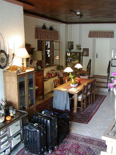 Een gezellige ruimte met een eetgedeelte, een grote houten tafel en verschillende lampen. Op de achtergrond bevinden zich ramen en trappen, terwijl bagagestukken op de vloer staan.