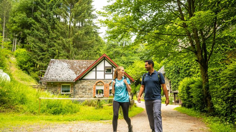 Ein Paar wandert auf einem Waldweg in der Natur. Im Hintergrund ist ein charmantes Haus zwischen Bäumen sichtbar.