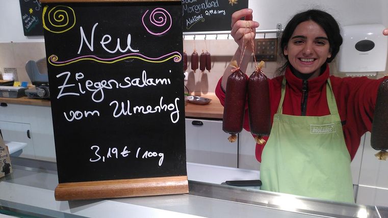 A woman is standing behind a counter and holding two sausages in her hand. Next to her is a sign that says "New: Goat Salami from Ulmenhof."