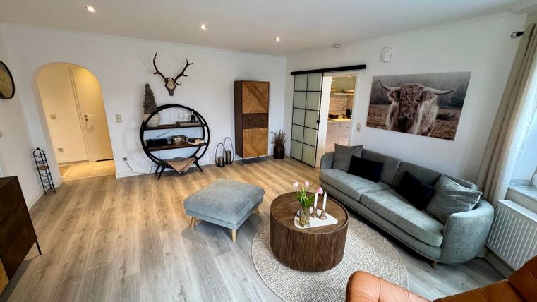 Ein modernes, helles Wohnzimmer mit gemütlicher Couch und Smart-TV, sowie einem Lesesessel und direktem Zugang zur Sonnenterrasse.