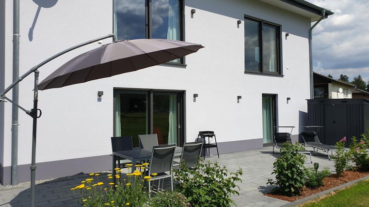 Ein modernes Haus mit großen Fenstern und einer Terrasse. Auf der Terrasse stehen Gartenmöbel und umgeben sind sie von bunten Blumen.