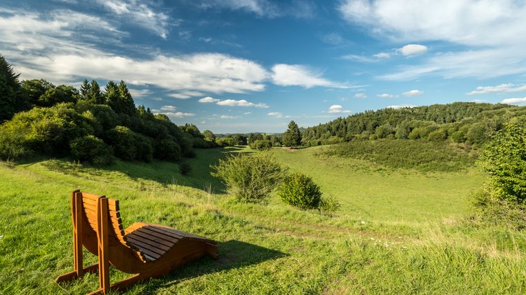 Holzliege auf einer Wiese mit Blick auf eine grüne Landschaft und blauen Himmel.