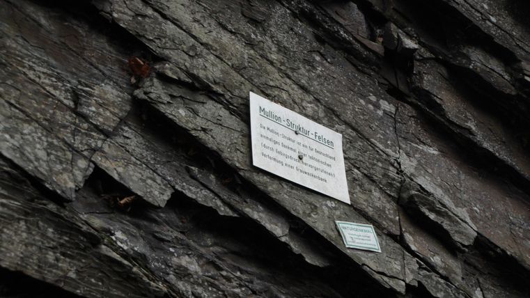 Eine steinige Wand mit einer Tafel, auf der Informationen stehen. Die Oberfläche ist rau und dunkel.