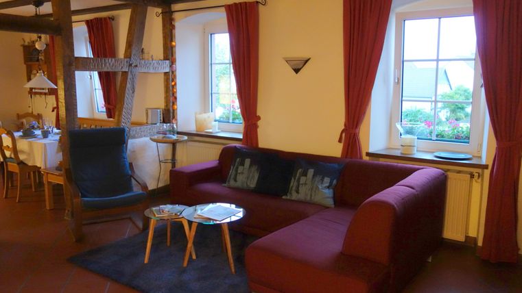 Een gezellige woonkamer met een rode bank en een zwart tapijt. Grote ramen met rode gordijnen zorgen voor veel daglicht.