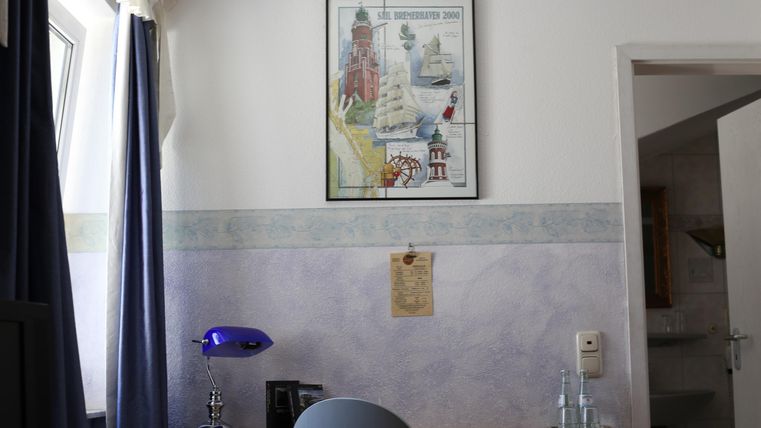 Ein heller Raum mit einem Schreibtisch und einem blauen Stuhl. An der Wand hängt ein Poster mit maritimem Motiv.