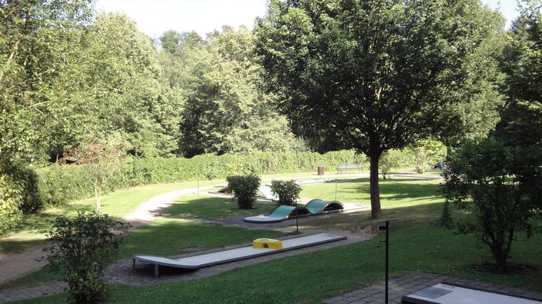 Een rustige park met groene bomen en een kronkelige pad. Op de voorgrond zijn ligbedden en een klein pad te zien.