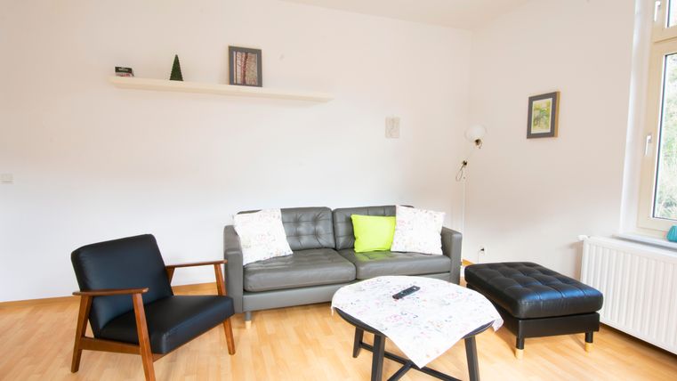 Ein modernes Wohnzimmer mit einem grauen Sofa, einem schwarzen Sessel und einem Tisch. Die Wände sind schlicht und hell, und es gibt ein Fenster mit natürlichem Licht.