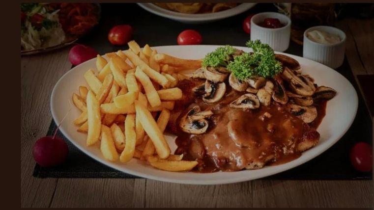 Ein Teller mit gebratenem Fleisch, überzogen mit einer Sauce und Pilzen. Dazu gibt es knusprige Pommes frites und frische Beilagen.