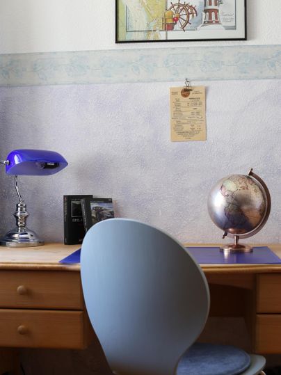 Ein Schreibtisch mit einer blauen Lampe, einem Globus und einem Bilderrahmen. Die Wand ist hell und zeigt dekorative Elemente.