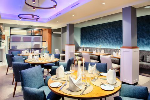 Modern restaurant met blauwe stoelen, ronde houten tafels en stijlvolle verlichting. De tafels zijn elegant gedekt.