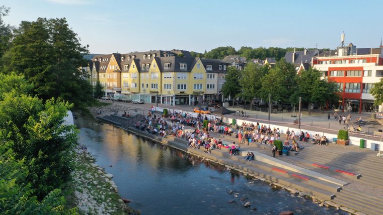 A lively riverside promenade with many people. In the background, colorful buildings and trees can be seen.