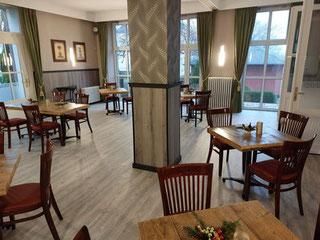 Ein gemütliches Restaurant mit Holzstühlen und Tischen. Große Fenster sorgen für viel Tageslicht und eine einladende Atmosphäre.