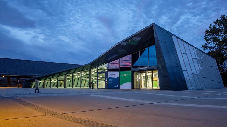 A modern building with distinctive, slanted architecture. It is illuminated in the evening, and the sky is partly cloudy.