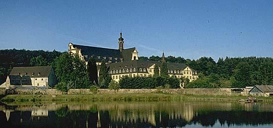 Ein malerisches Klostergebäude am Ufer eines ruhigen Sees, umgeben von grünen Wäldern. Die klare Reflexion der Architektur im Wasser verstärkt die Schönheit der Umgebung.