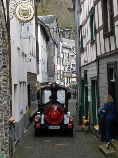 A narrow, charming alley with historic buildings and a colorful train passing through the street. In the background, a person can be seen observing the surroundings.