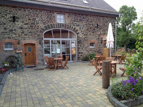 Ein gemütlicher Außenbereich mit Holztischen und Stühlen vor einem alten Steinhaus. Einige Pflanzen und eine Sonnenschirm sorgen für eine einladende Atmosphäre.