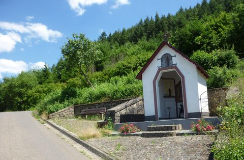 Eine kleine Kapelle steht an einem schmalen Weg, umgeben von grünen Bäumen. Der Himmel ist klar und blau.