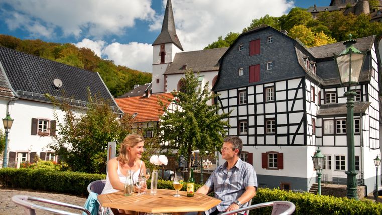 Ein Paar sitzt entspannt an einem Tisch im Freien mit Getränken. Im Hintergrund sind charmante Fachwerkhäuser und ein blauer Himmel mit Wolken zu sehen.