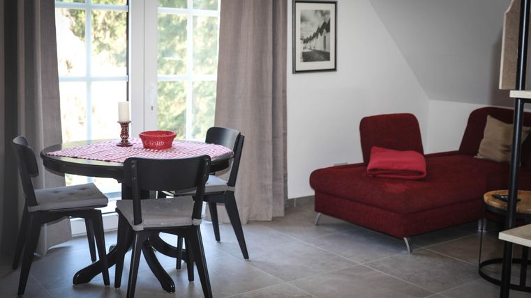 A cozy living area with a round table, chairs, and a red sofa. Large windows let in a lot of light and create an inviting atmosphere.