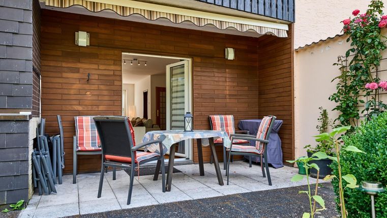 Eine gemütliche Terrasse mit einem Tisch und Stühlen. Im Hintergrund ist ein Zugang zum Haus sowie Pflanzen und Rosen zu sehen.