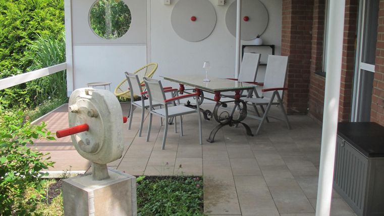 Eine Terrasse mit einem Tisch und Stühlen, umgeben von grünen Pflanzen. An der Wand befindet sich eine dekorative Skulptur.
