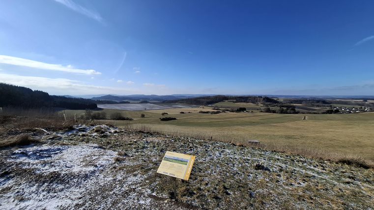 A vast landscape with gentle hills and a clear sky. In the foreground, there is an information board.