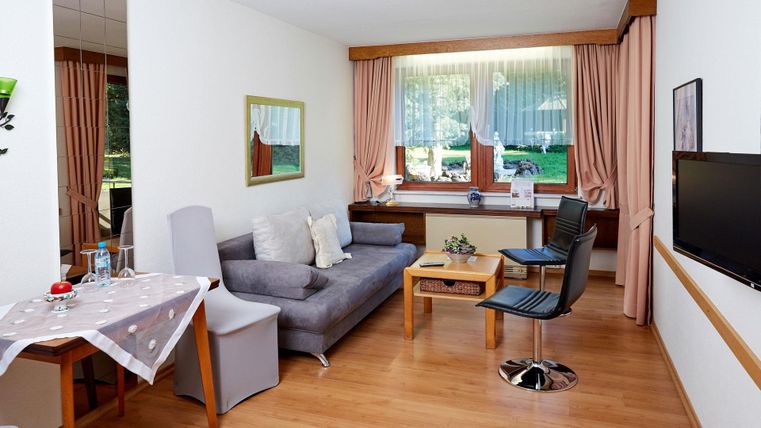 Een gezellige woonruimte met een grijze bank, een tafel en een stoel. Grote ramen zorgen voor veel daglicht en uitzicht op de buitenlucht.