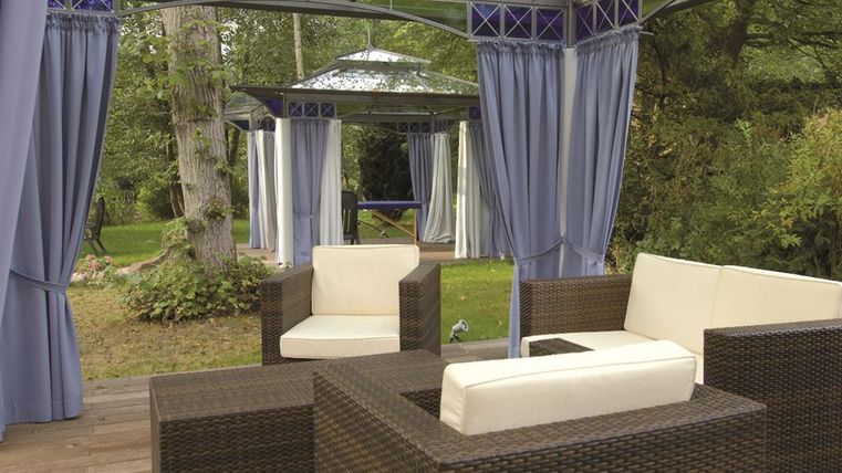 A cozy outdoor area with rattan furniture and bright cushions. Surrounded by trees and curtains, it offers a relaxed atmosphere.