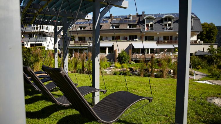 A cozy lounge area under a canopy in a beautiful garden. In the background, modern buildings and green spaces are visible.