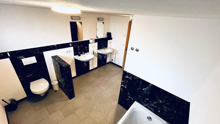 A modern bathroom with elegant black walls and bright tiles. There is a bathtub, two sinks, and a toilet.