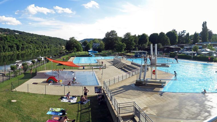 A swimming pool with multiple basins and many visitors. The surroundings are green and there are play equipment for children.