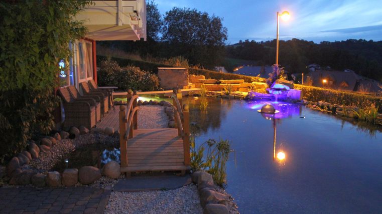 Ein schöner Garten mit einem Teich und einer Holzbrücke. Die Umgebung ist abends sanft beleuchtet.