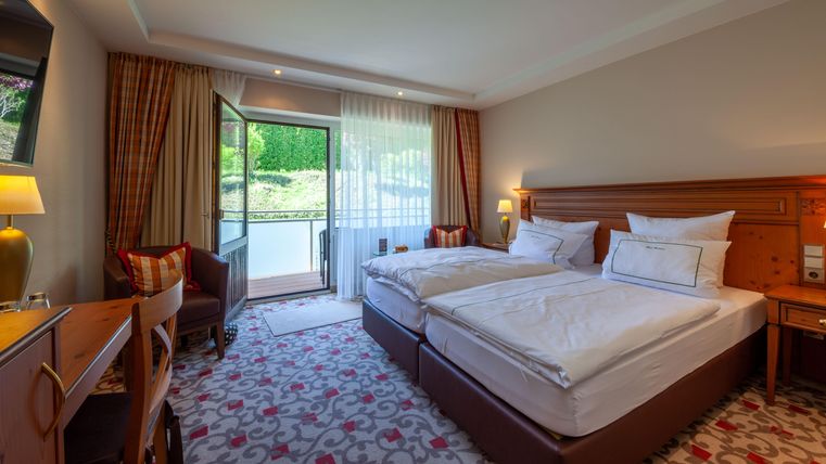 Ein modernes Hotelzimmer mit zwei Einzelbetten, einem Schreibtisch und einem Zugang zu einem Balkon. Große Fenster lassen viel Tageslicht herein und bieten einen Blick ins Grüne.