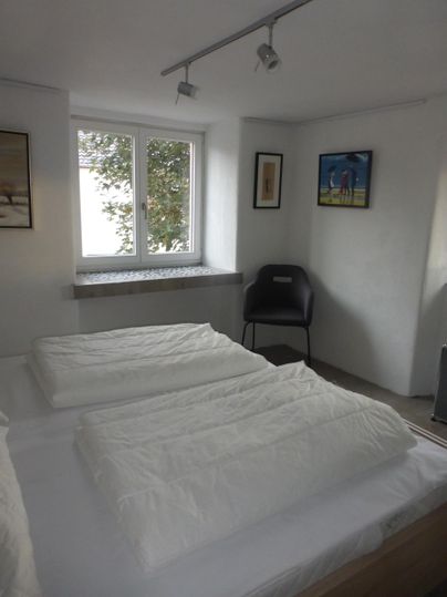 Ein heller, moderner Schlafzimmer mit zwei Betten und frischen weißen Bettdecken. Im Raum gibt es ein Fenster mit Blick nach draußen und einige Wandbilder.
