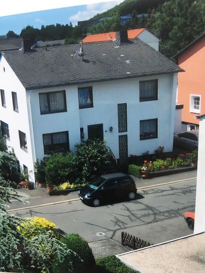 Een wit woonhuis met luiken en een groene voortuin. Op straat staat een zwarte auto geparkeerd.