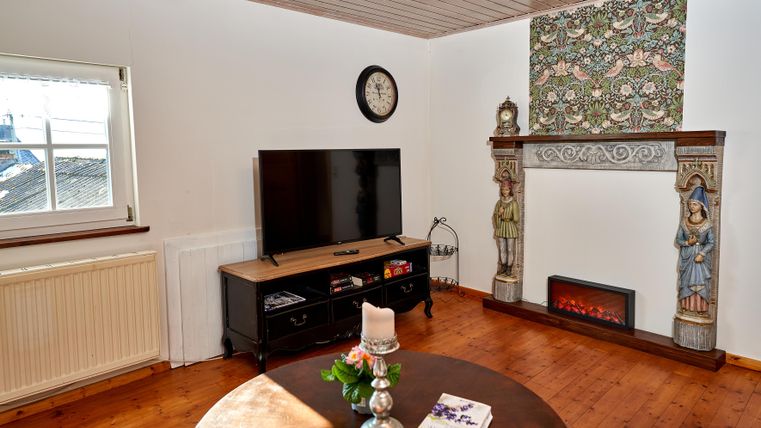 A cozy living room with a television and a decorative fireplace. On the wall is an artistically designed wall covering and a round table with a flower vase.
