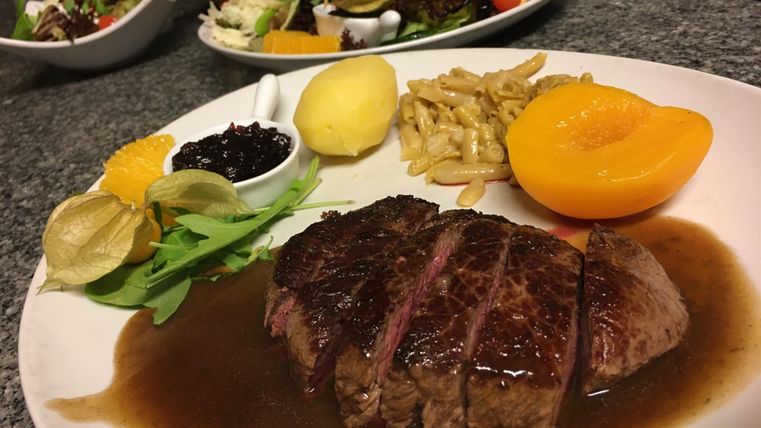 Ein Teller mit saftigem Steak in dunkler Soße, serviert mit Kartoffeln, Pfirsich und verschiedenen Beilagen. Im Hintergrund sind frische Salate zu sehen.