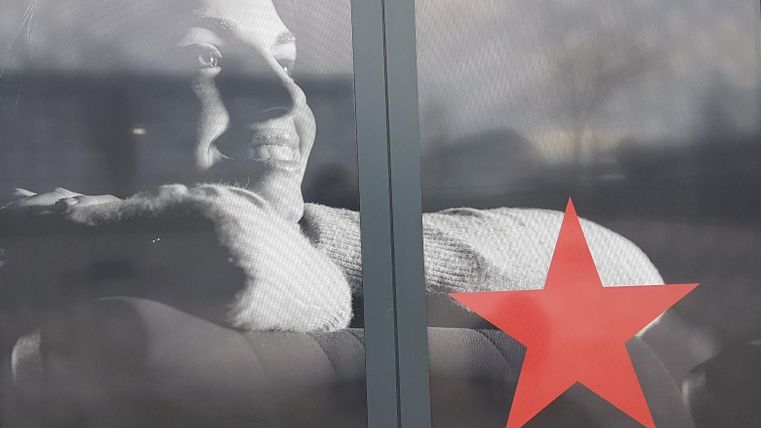 Ein Porträt einer nachdenklichen Person, die entspannt mit dem Kopf auf den Händen liegt. Im Vordergrund befindet sich ein rot star auf der Glasfläche.