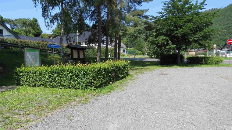 Eine ruhige Straße mit Kiesbelag, umgeben von Bäumen und Büschen. Im Hintergrund sind einige Gebäude und ein Spielplatz zu sehen.