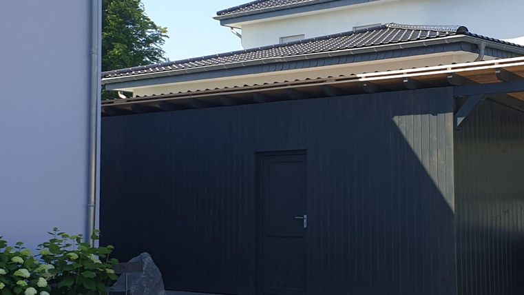 Ein modernes, schwarzes Gartenhaus steht neben einem Wohnhaus. Im Vordergrund sind grüne Pflanzen und ein kleiner Stein sichtbar.