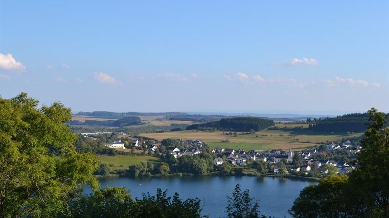 Eine malerische Landschaft mit einem See und grünen Hügeln. Im Hintergrund sind kleine Häuser und ein klarer blauer Himmel zu sehen.