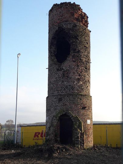Een oude, beschadigde bakstenen toren staat in een open landschap. Het bovenste deel is zwaar vervallen en de lucht op de achtergrond is helder.