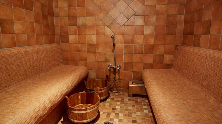Sauna room with tiled benches, wooden buckets, and shower. The walls and floor are covered with brown tiles.