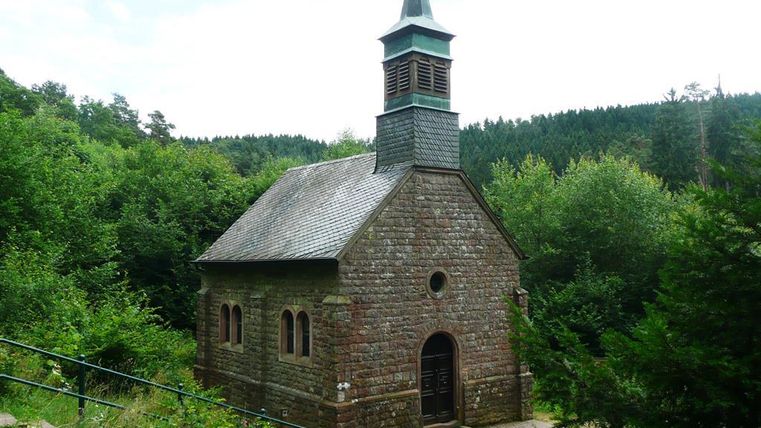 Kleine steinerne Kapelle mit spitzem Turm, umgeben von dichtem Wald. Der Eingang ist von Pflanzen umgeben.