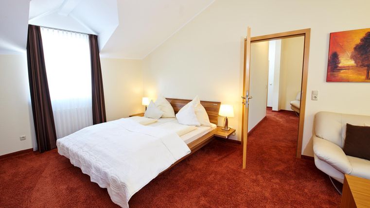 Ein helles Schlafzimmer mit einem großen Bett, weich beleuchtet von zwei Nachttischen. Der Raum hat einen gemütlichen Teppich und einen Zugang zu einem weiteren Raum.